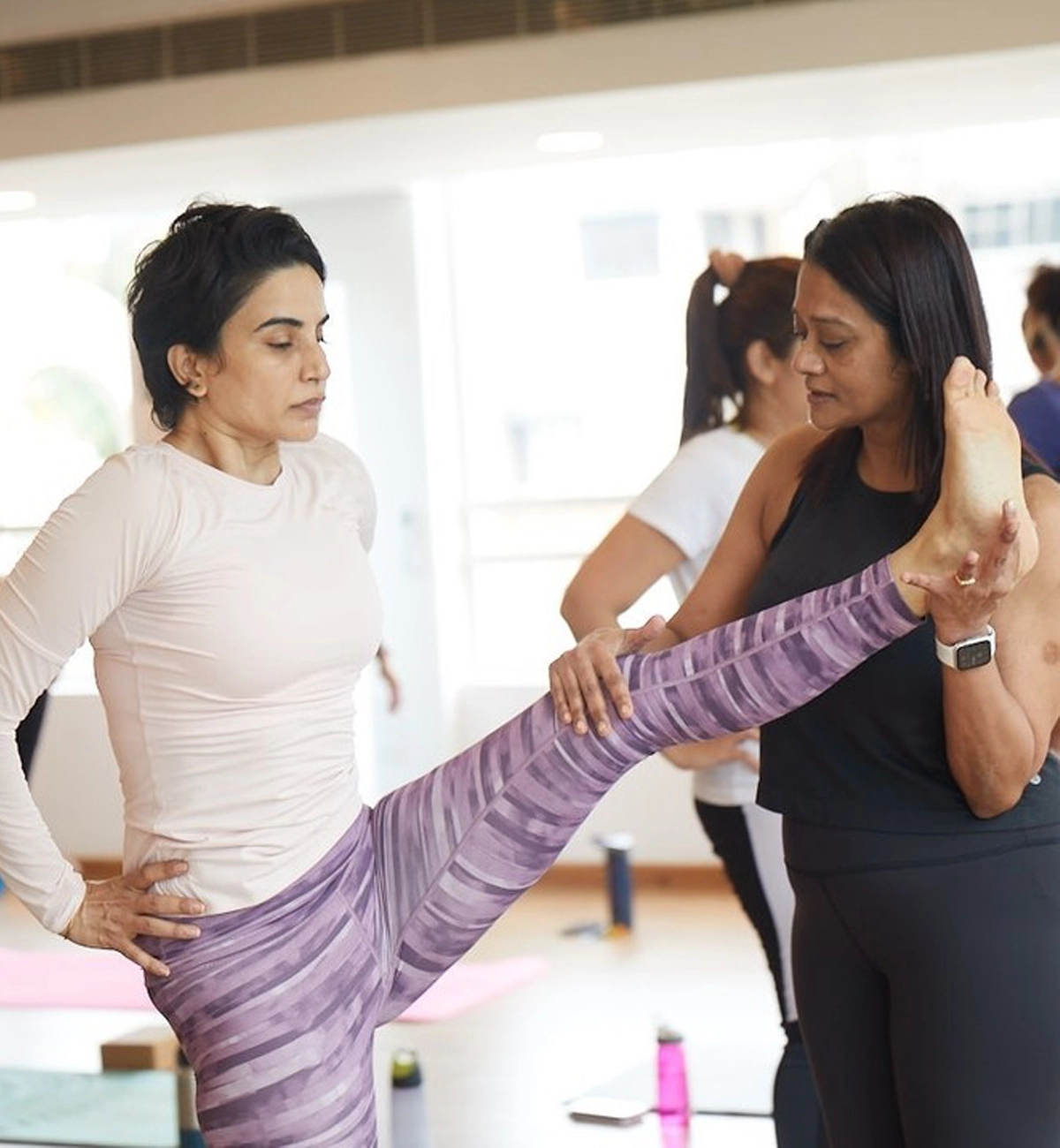 A yoga instructor helping a woman stretch her leg during a class.